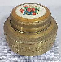 Side and top view of vintage musical powder box showing floral ceramic inset on lid and embossed gold-tone base.
