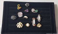 12 vintage costume jewelry rings arranged in black display tray showing varied designs and color tones.