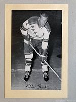 Front view of black and white hockey photo of Eddie Shack in New York Rangers uniform, adhered to cream cardboard backing.