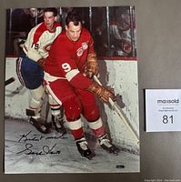 Full color photo showing Gordie Howe in a red hockey uniform skating on the ice with an opponent, signed in black Sharpie.