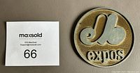 Top view of the Montreal Expos pewter drink coaster beside a MaxSold card for size reference, showing logo and text detail.