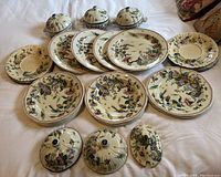 Full lot view showing arrangement of dinner plates, bowls, lids, and bases on a cloth surface. Visible intricate painted designs with floral and bird themes.