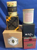 Photo showing six assorted scented candles, five in their original decorative boxes, one rolled beeswax candle wrapped with MacKenzie-Childs band.
