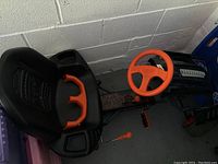 Side view of the black seat with orange handles, foot pedals, and part of the steering wheel visible next to a white wall.