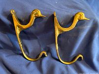 Two brass duck wall hangers side by side on a blue fabric background showing the duck head and hook shape.