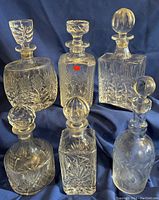 All six decanters arranged on blue fabric background showing different shaped lids and intricate cut glass designs.