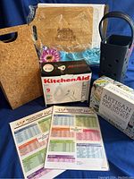 Photo of all lot contents together showing cutting boards, boxed KitchenAid mixer, barbecue sauce kit, pressure cooker time charts, and blue wine bottle holder.
