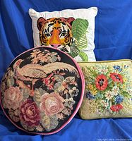 Three pillows on blue fabric background: square tiger crewel pillow, round bird needlepoint pillow with pink trim, rectangular floral needlepoint pillow
