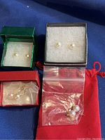 Top view of four sets of pearl earrings displayed in small boxes and a plastic bag on a dark blue fabric background.
