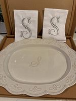 Photo showing white porcelain platter with beaded decorative border and embossed 'S' monogram in center, placed inside an open original brown cardboard box with two white cloth napkins visible.
