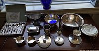 General view of various silver and silver-plated items arranged on wooden surface showing multiple bowls, cups, serving pieces, and silverware box.
