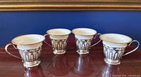 Four demitasse cups with sterling silver bases and white porcelain liners with gold trim shown from the front on a reddish surface