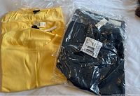 Photo showing a yellow charming cardigan and shell sweater, fully out of packaging, with original tags attached, lying next to a black charming cardigan and shell set mostly inside plastic packaging with tags visible.