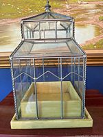 Front view showing square terrarium with glass panes framed in dark metal with decorative twists and zigzag pattern on each side with beige tray base inside.