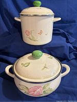 Two ceramic pans with lids showing floral and insect designs, sitting on blue cloth backdrop.
