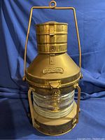 Full frontal view of the brass anchor oil lamp showing the top cap, hanging handle, Fresnel lens glass, and solid brass body with embossed ANCHOR brand.