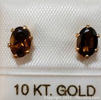 Close-up photo of the pair of oval dark brown smokey quartz earrings set in 10 karat yellow gold with six prongs each, placed on fabric stamped '10 KT GOLD'.