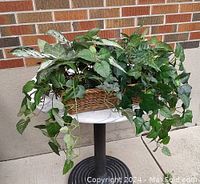 Front view of faux plant display in wicker basket on small round table showing dense foliage and leaf variegation.