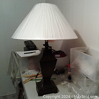 Full view of the brown all metal lamp standing on a table showing its base, cord, and white pleated shade.