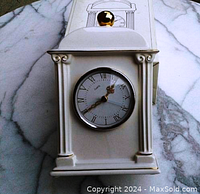 White ceramic mantle clock with gold trim, showing front clock face and gold spherical finial on top. Clock features Roman numerals and gold-colored hands. Clock is placed on white marble surface with original box behind it.