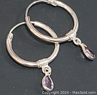 Close-up photo showing pair of sterling silver hoops with small dangling teardrop amethyst gemstones against dark background.