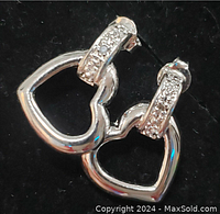 Pair of sterling silver open heart-shaped earrings with diamond-studded rectangular bars on black background.