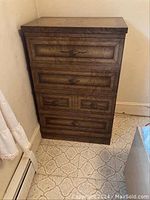 Front view of a tall wooden chest of drawers with five drawers, standing against a wall on carpet flooring.