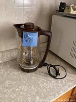 Electric glass coffee pot with floral gold design, brown handle, lid, power base and electric cord on countertop