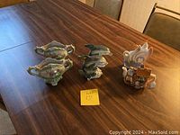 Six decorative knickknacks including floral vessels, dolphin figurines, swan vase, and cottage teapot figurine arranged on wooden table