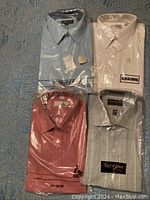 Four men's dress shirts, new with tags, folded and sealed in plastic, showing different colors and brands.