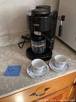 Photo showing black Saillon espresso maker with glass carafe and two Lynns Fine China espresso cups and saucers with blue floral design placed on counter.