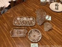 Five glass items on wooden table: water pitcher, milk pitcher, two serving dishes, and ashtray.
