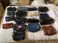 Lot of 17 handbags and purses laid out on a bed showing variety of styles, sizes, and colors mostly black with one brown item.