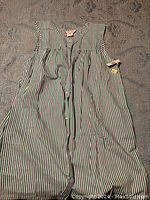 Full view of one sleeveless house dress with vertical stripes in green, purple, and white colors laid flat showing button front and tie waist.