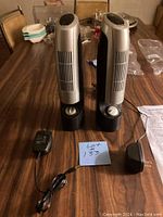 Two ionic mini air purifiers standing upright on a wooden table with two 12V power adapters and a lot identification card.