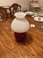 Side angled view of kerosene lamp showing hobnail white milk glass shade and red glass base on wooden table.