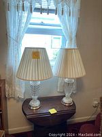 Pair of glass and metal living room table lamps with pleated cream lampshades placed on a wooden table under a window.