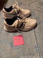 Side view of one Nike white sneaker with black accents placed on a patterned surface, next to a lot sticker.