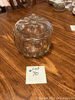 Clear glass cookie jar shaped like a pumpkin on a wooden table with a lot number note.