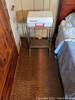 Record player closed case on metal stand between wooden furniture and bed, illustrating size and placement context.