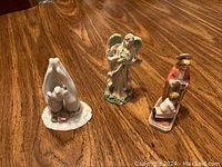 Three religious figurines on a wooden table: praying hands, angel, and saint with kneeling child.