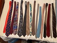 Top view of 16 men's ties laid flat in a row showing various colors and patterns, including polka dots, floral, solid, and striped designs.