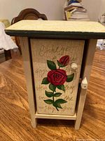 Front view of small wooden cabinet with painted red rose and white knob on the door.