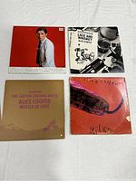 Four Alice Cooper record covers laid out on white cloth: Muscle of Love in cardboard sleeve, Lace and Whiskey black and white art, Killer cover with purple tail image, and an album with man in white jacket.