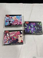 Three boxed jigsaw puzzles laid out on fabric, showing front colorful anime-style character art from Hazbin Hotel