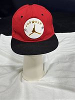 Front view of red Jordan cap showing circular white patch with gold Jumpman logo, JORDAN text and star, black brim