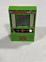 Front view of the green Konami Frogger handheld arcade game powered off, showing Frogger logo, frog decals, joystick, buttons, and speaker grills.