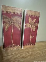 Photo showing two vertical canvas prints with tropical plant motifs on red backgrounds placed against a neutral wall and on a wooden surface.