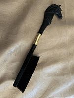 Full length of antique horse brush showing black and gold coloring, with horse head handle and rectangular black bristle brush.