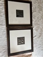 Two framed antique metal Fleur de Lys embossed panels. Each panel is centered in a white matte and dark brown wooden frame, showing oxidation and patina on the metal.